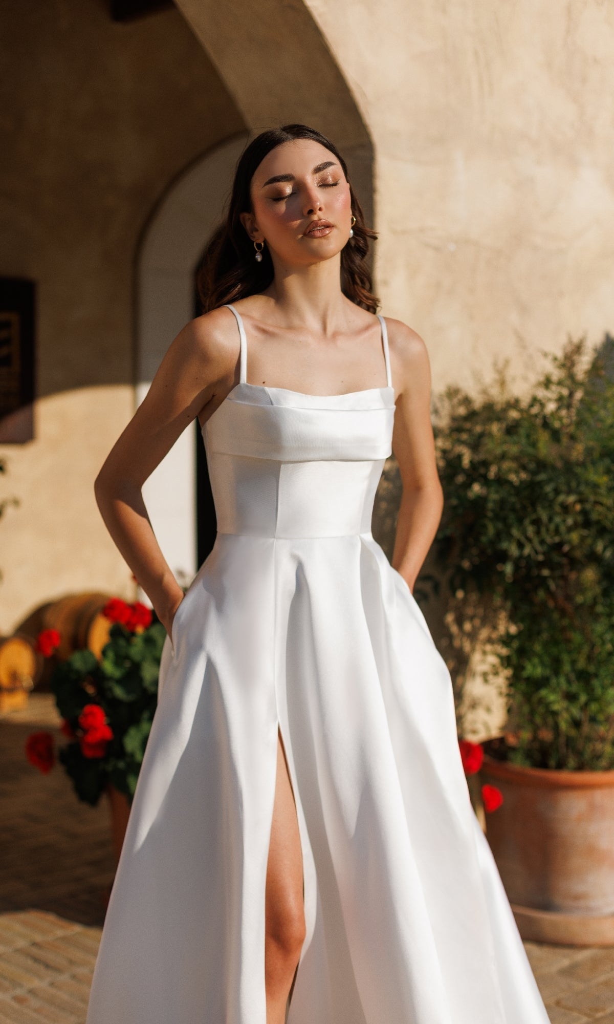 A woman stands outdoors in sunlight among plants and flowers, wearing the Tina Holly TE305 Classic Long White Formal Dress—a sleeveless, a-line gown with a thigh-high slit.
